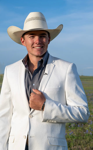 Man wearing a white cowboy hat and light-colored suit standing outdoors with a clear sky.