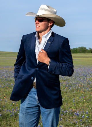 Man wearing a navy blazer, white shirt, and cowboy hat standing in a field of blue flowers.